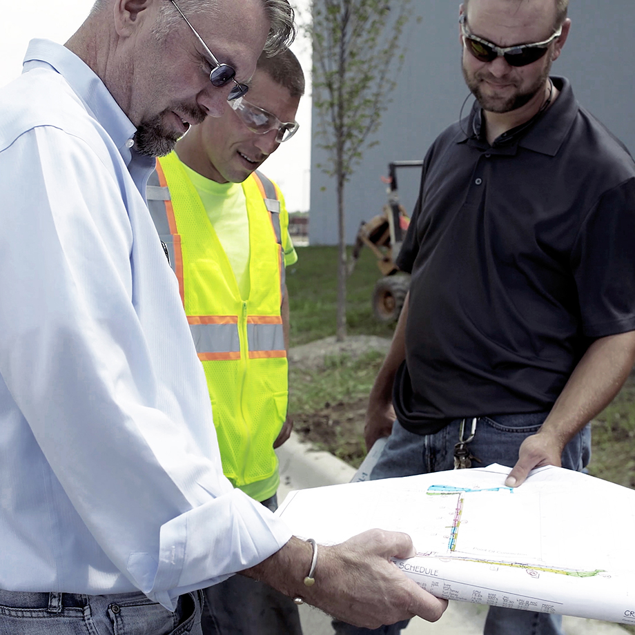 Three construction professionals, one in a high-visibility vest, review building plans on a worksite with construction machinery in the background.